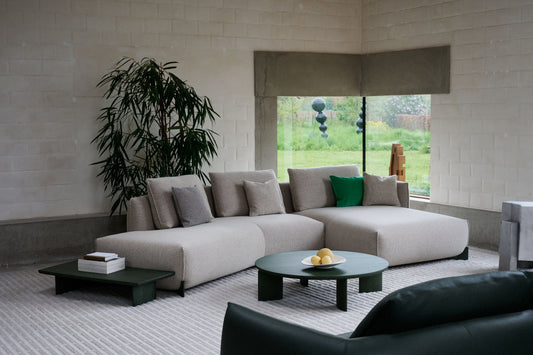 A contemporary beige fabric Naila sectional sofa by Leolux, featuring a smooth and sleek design with integrated side table, displayed in a living room setting with decorative cushions, tables, and a large window in the background.