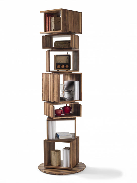 A tall, modern wooden rotating bookshelf made by Riva 1920 with multiple open cubby shelves, made of solid wood  styled with books, a small radio, cups, and other décor objects, set against a white background.